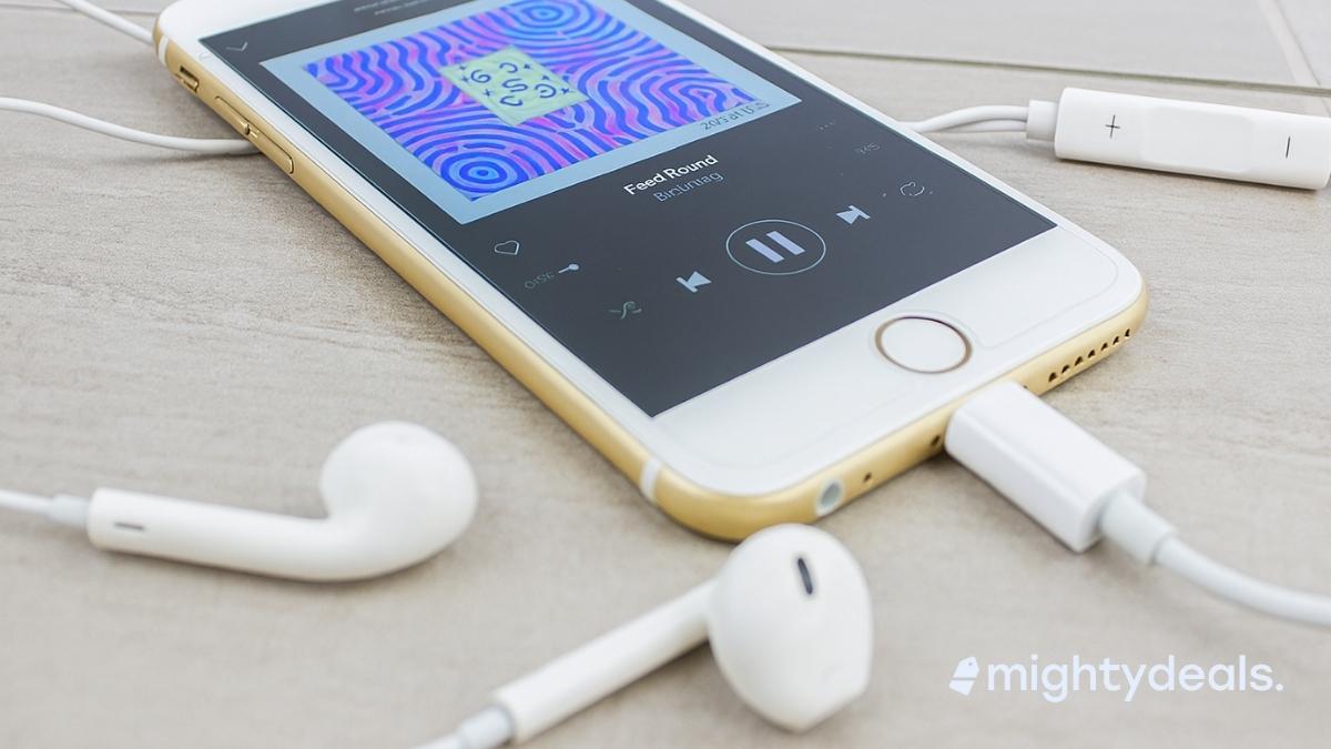 A gold iPhone lies on a light gray tiled surface with its screen displaying a music player app and colorful album art. White wired EarPods are plugged into the phone, with the earbuds and in-line remote resting nearby.