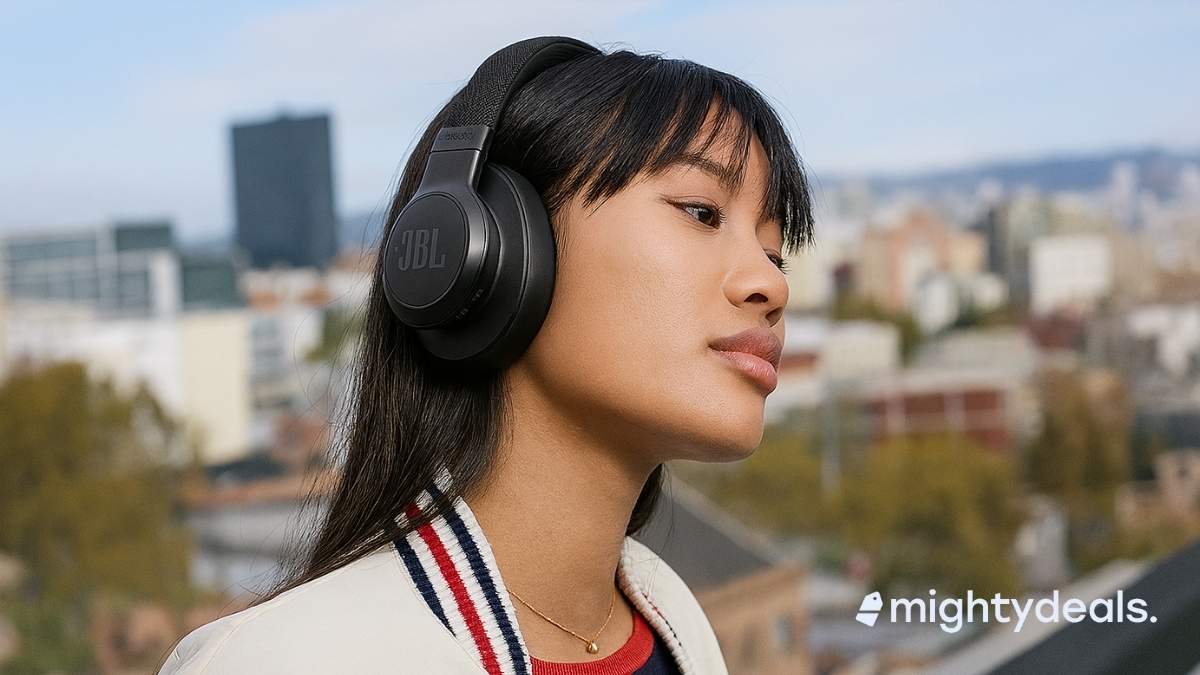 A young Asian woman wearing black JBL over-ear headphones stands on a rooftop, gazing thoughtfully into the distance. She has straight black hair with bangs and is dressed in a cream varsity jacket with red and blue stripes over a navy top. The blurred background shows a cityscape with buildings, trees, and a blue sky.
