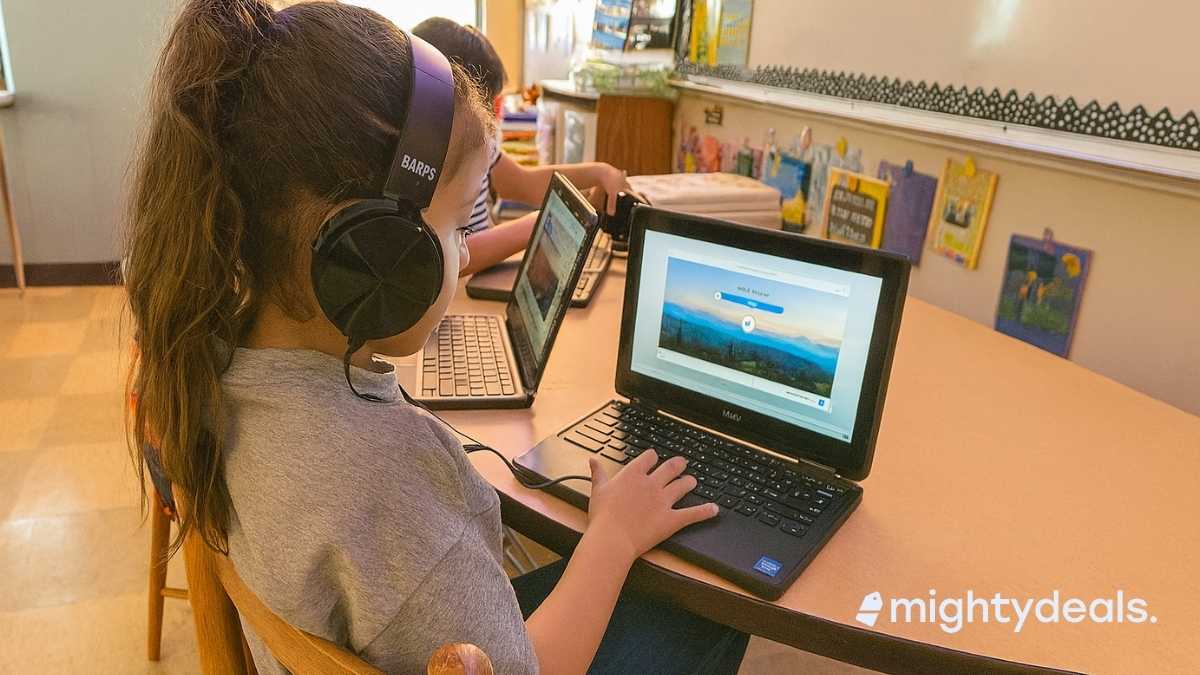 A young girl wearing over-ear headphones sits at a small wooden desk in a classroom, focused on a laptop screen. Other students are working in the background, and colorful artwork decorates the classroom walls.