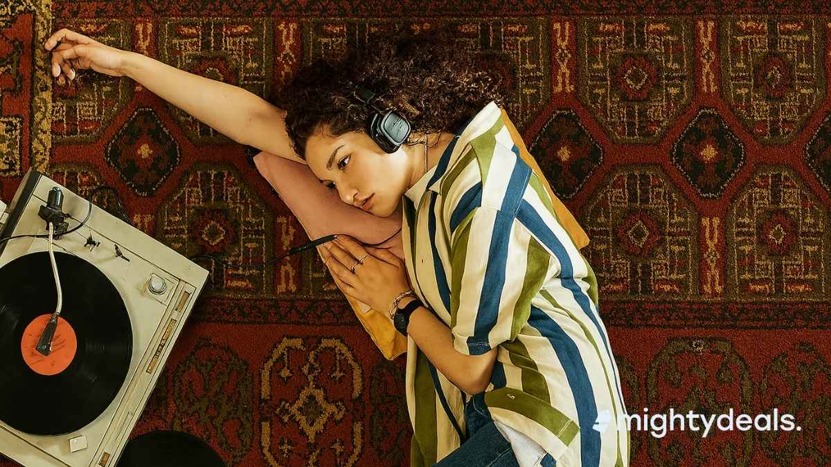 A young woman with curly hair lies on a patterned red rug, wearing black over-ear headphones and resting her head on a pillow. She gazes toward a spinning vinyl record on a vintage turntable placed beside her, with another record and an open notebook nearby. She wears a striped shirt and ripped jeans, creating a cozy and nostalgic atmosphere.