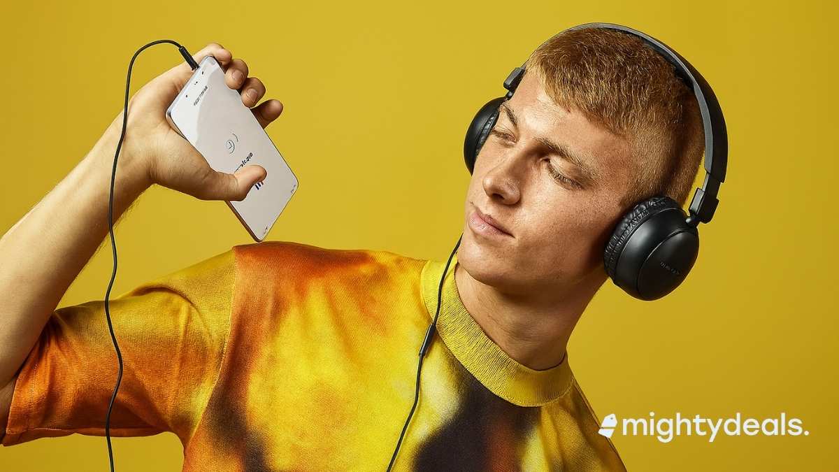 A young man wearing black over-ear headphones looks at a white smartphone connected via a wired USB-C cable. He is dressed in a vibrant tie-dye shirt with orange and yellow tones, set against a solid yellow background.