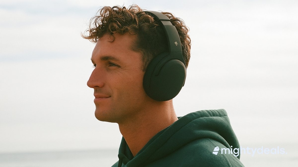 A young man with curly brown hair wears black over-ear Skullcandy headphones and a green hoodie while standing near the ocean, gazing calmly into the distance.